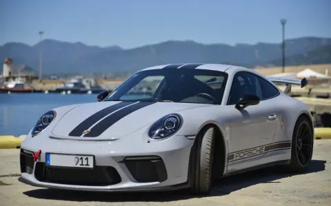 Szary Porsche 911 z czarnymi pasami, stojący nad wodą. Zastanawiasz się nad porsche 911 cena?