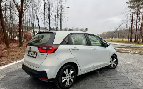 Biały Honda Fit stoi na mokrej, brukowanej nawierzchni. W tle drzewa i budynki.