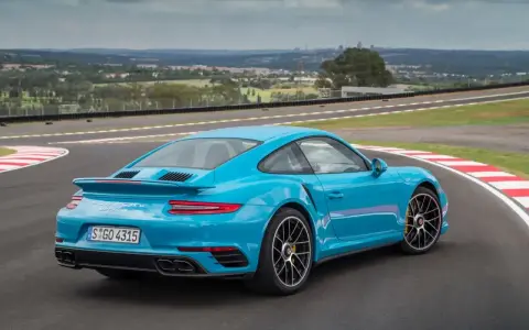 Błękitne Porsche 911 Turbo S na torze wyścigowym, z widokiem na miasto w tle.