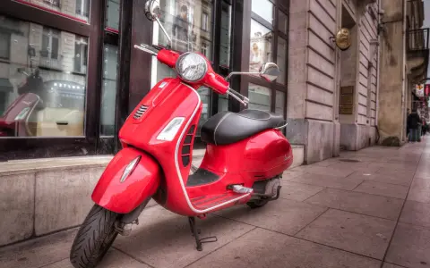Czerwony skuter Vespa, idealny do zdobycia prawa jazdy AM, stoi na chodniku przed budynkiem.