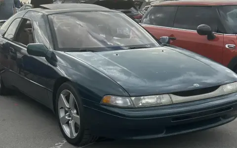 Subaru SVX w ciemnozielonym kolorze, z charakterystycznymi oknami w stylu "glass-canopy", zaparkowany obok czerwonego samochodu.
