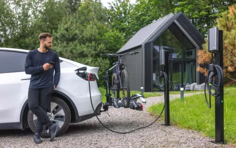 Mężczyzna z telefonem przy białym samochodzie elektrycznym, który ładuje się obok stojaka z rowerami. Obok stoją dwie ładowarki do samochodów elektrycznych.