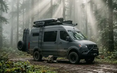 Szary bus z napędem 4x4, gotowy na przygodę w lesie. Na dachu bagażnik z wyposażeniem, z boku drabinka i koło zapasowe.