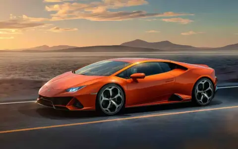 Pomarańczowy Lamborghini Huracan pędzi po pustej drodze nad jeziorem o zachodzie słońca.