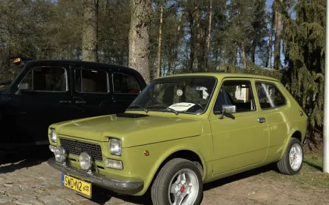 Zielony fiat 127 z lat 70. stoi obok czarnej taksówki na tle drzew.