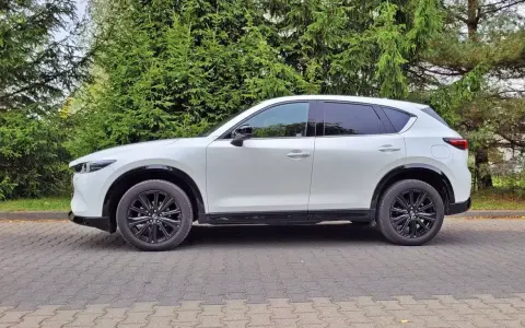 Nowa Mazda CX-5 w białym kolorze, zaparkowana na brukowanym podjeździe, otoczona zielenią drzew.