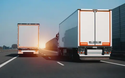 Dwa białe tiry jadą autostradą. Na jednym z nich widać znak zakazu wyprzedzania dla ciężarówek.
