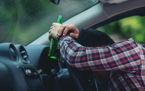 Mężczyzna w kratę trzyma butelkę, opierając głowę na kierownicy. To obraz ostrzegawczy przed jazdą pod wpływem alkoholu, przypominający o zagrożeniach i potrzebie blokady alkoholowej.