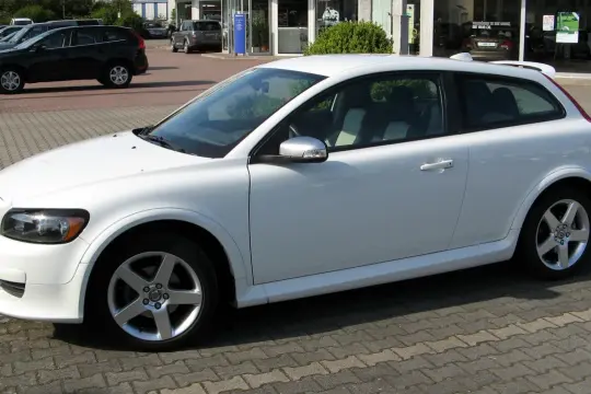 Białe Volvo C30 z felgami pięcioramiennymi stoi na parkingu przed salonem samochodowym.