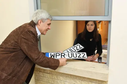 Mężczyzna odbiera nowe tablice rejestracyjne od pracownicy urzędu.