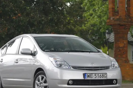 Srebrny Toyota Prius Plus z tablicą WE 5168G zaparkowany obok ceglanej budowli i drzew.