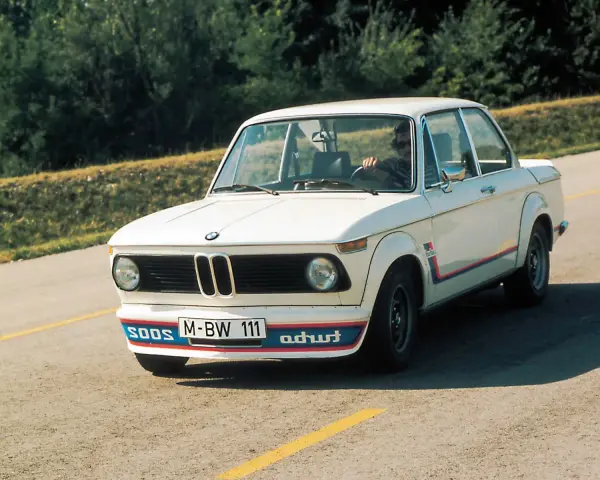 Białe BMW E20 z kolorowymi pasami i napisem "turbo" na błotniku, z kierowcą za kierownicą, jedzie po asfaltowej drodze.