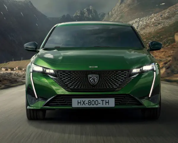 Zielony Peugeot 308 T7 jedzie drogą w malowniczym, górskim krajobrazie z widokiem na małą wioskę nad wodą.