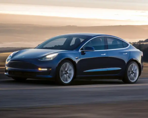 Niebieska Tesla pędzi po drodze, osiągając prędkość do 100 km/h. Tło rozmywa się w złotej poświacie zachodzącego słońca.