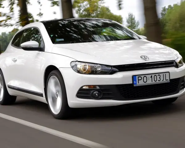 Biały Volkswagen Scirocco pędzi po drodze wśród drzew.