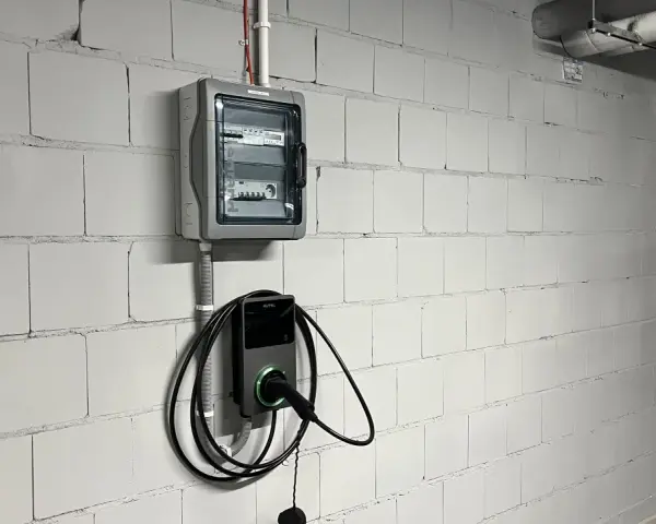 Nowoczesne gniazdo do ładowania samochodu elektrycznego Autel zamontowane na białej, ceglanej ścianie obok skrzynki z bezpiecznikami.