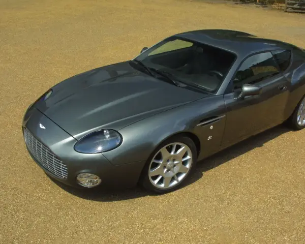 Szary Aston Martin DB9 na żwirowym podjeździe. Zastanawiasz się, ile kosztuje Aston Martin? To auto to marzenie wielu.
