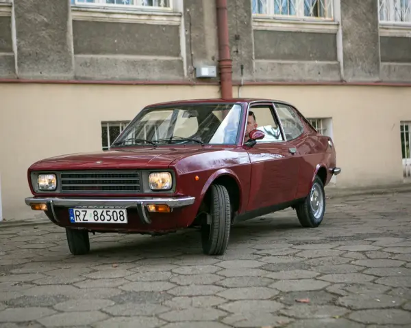 Czerwony fiat 128, klasyk z lat 70., zaparkowany na brukowanym dziedzińcu przed budynkiem.
