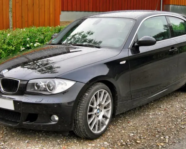 Czarny BMW e81, lśniący po deszczu, stoi na żwirowej drodze obok zielonego żywopłotu i drewnianego ogrodzenia.
