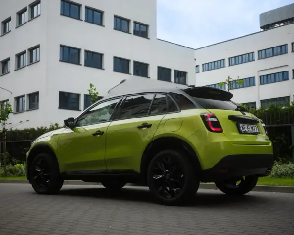 Jaskrawozielony Fiat 600 z czarnymi dodatkami parkuje przed nowoczesnym budynkiem.