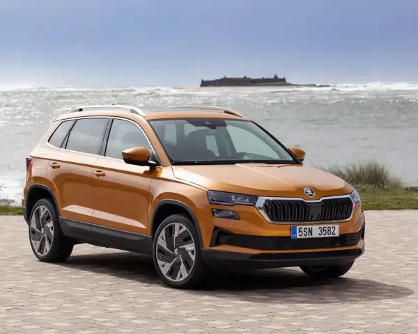 Pomarańczowa Skoda Karoq na tle morza i zamku. Sprawdź aktualny skoda cennik i wybierz swój wymarzony model.