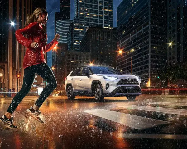 Biegaczka w deszczowy wieczór w mieście, obok niej biała Toyota RAV4 Hybrid. Zastanawiasz się, ile pali Toyota RAV4 Hybrid?