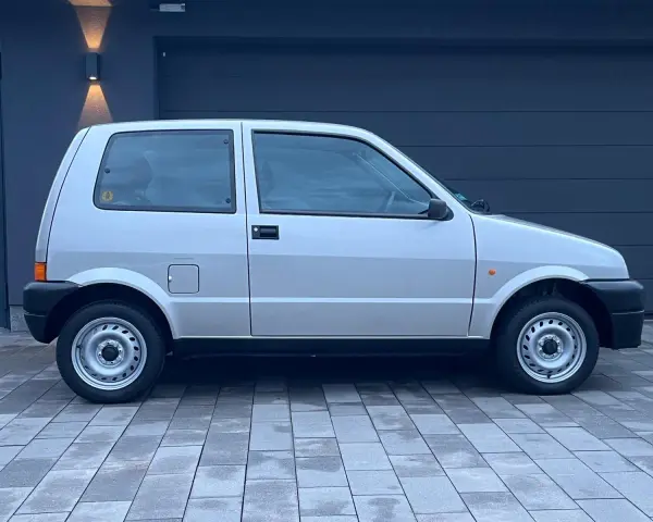 Srebrny Fiat Cinquecento na tle ciemnego garażu, z charakterystycznymi felgami i czarnymi zderzakami.