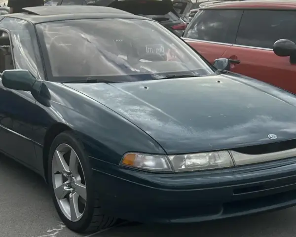 Subaru SVX w ciemnozielonym kolorze, z charakterystycznymi oknami w stylu "glass-canopy", zaparkowany obok czerwonego samochodu.