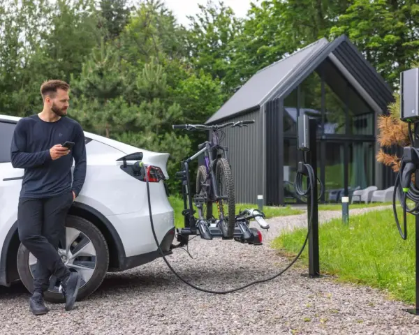 Mężczyzna z telefonem przy białym samochodzie elektrycznym, który ładuje się obok stojaka z rowerami. Obok stoją dwie ładowarki do samochodów elektrycznych.