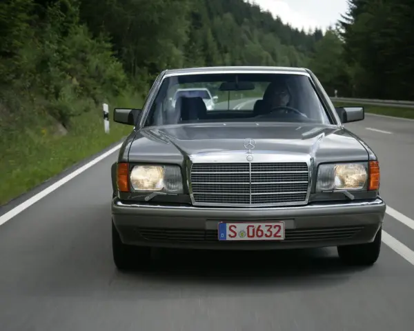 Szary Mercedes W126 jedzie drogą wśród zielonych lasów.