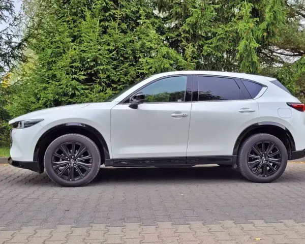 Nowa Mazda CX-5 w białym kolorze, zaparkowana na brukowanym podjeździe, otoczona zielenią drzew.