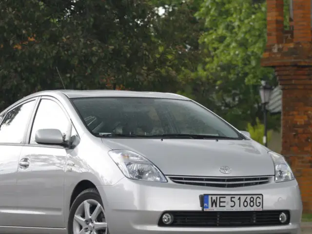 Srebrny Toyota Prius Plus z tablicą WE 5168G zaparkowany obok ceglanej budowli i drzew.