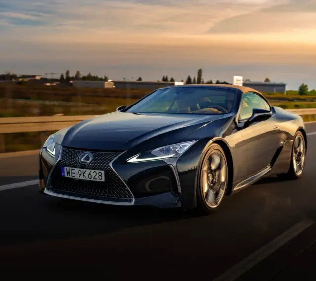 Ciemny Lexus LC 500 z beżowym dachem sunie po drodze podczas zachodu słońca. Sprawdź jego lexus lc 500 cena.