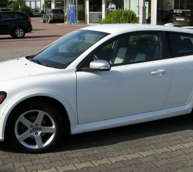 Białe Volvo C30 z felgami pięcioramiennymi stoi na parkingu przed salonem samochodowym.