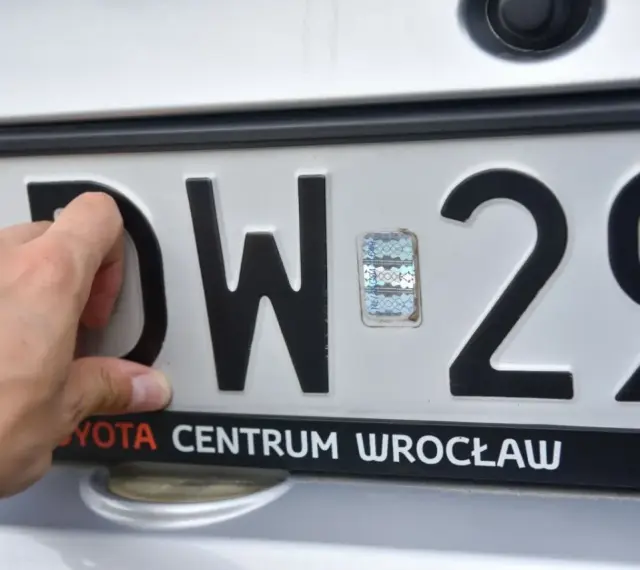Dłoń dotyka tablicy rejestracyjnej z literami DW i numerem 29. To tablice rejestracyjne województwa dolnośląskiego.
