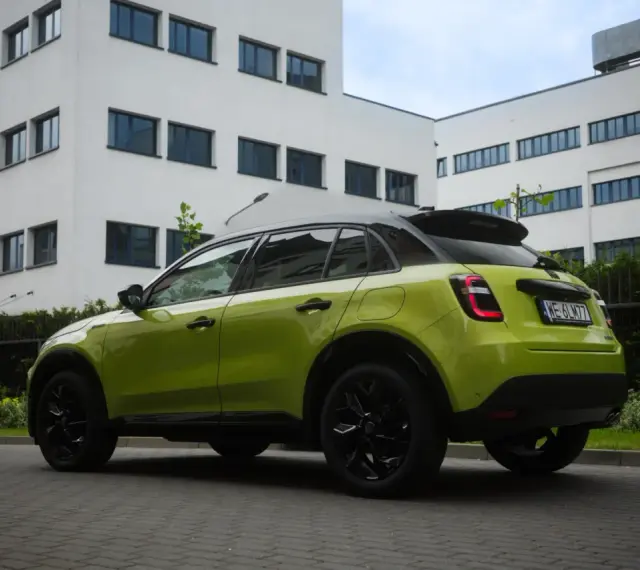 Jaskrawozielony Fiat 600 z czarnymi dodatkami parkuje przed nowoczesnym budynkiem.