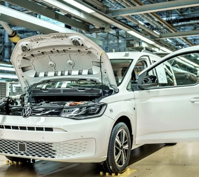 Biały samochód elektryczny na linii produkcyjnej. Widać otwartą maskę i drzwi, a także elementy silnika. Powstają tu polskie samochody.