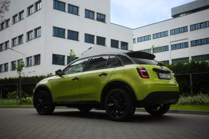 Jaskrawozielony Fiat 600 z czarnymi dodatkami parkuje przed nowoczesnym budynkiem.