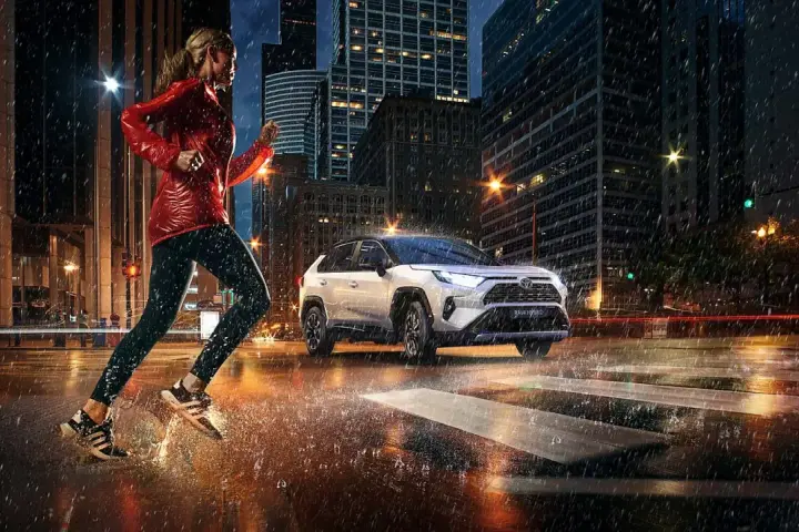Biegaczka w deszczowy wieczór w mieście, obok niej biała Toyota RAV4 Hybrid. Zastanawiasz się, ile pali Toyota RAV4 Hybrid?