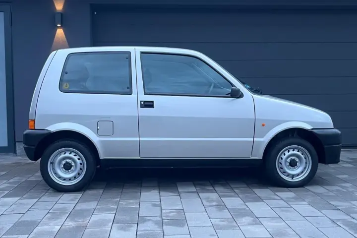 Srebrny Fiat Cinquecento na tle ciemnego garażu, z charakterystycznymi felgami i czarnymi zderzakami.