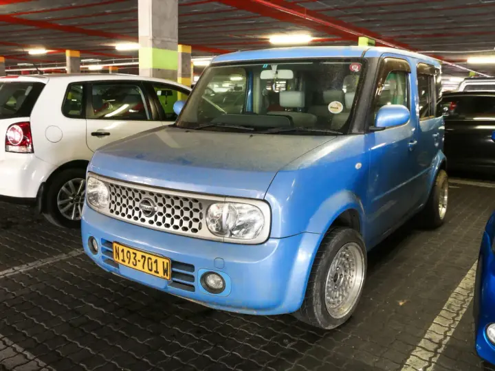 Niebieski Nissan Cube z charakterystycznym grillem i tablicą rejestracyjną N193-701 W, zaparkowany obok białego Volkswagena.