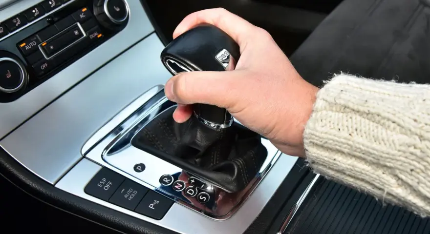 Dłoń trzyma gałkę zmiany biegów w samochodzie. Widoczna jest automatyczna skrzynia biegów DSG z przyciskami ESP, Auto Hold i czujnikami parkowania.