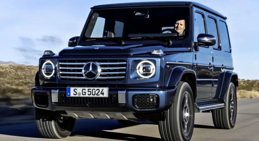 Ciemnoniebieski Mercedes G Klasa jedzie po pustynnej drodze. Kierowca uśmiecha się. Sprawdź mercedes g klasa cena!
