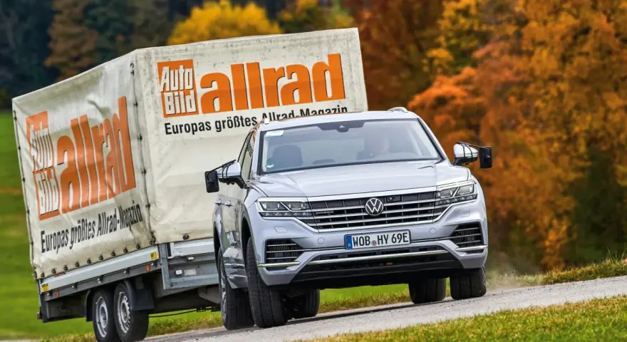 Srebrny Volkswagen Touareg holuje przyczepę z logo "Auto Bild allrad", jadąc po krętej drodze wśród jesiennych drzew.