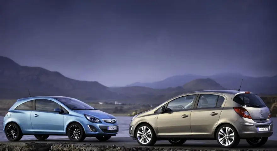 Dwa samochody Opel Corsa D, jeden niebieski, drugi beżowy, stoją na kamiennym murze nad wodą.