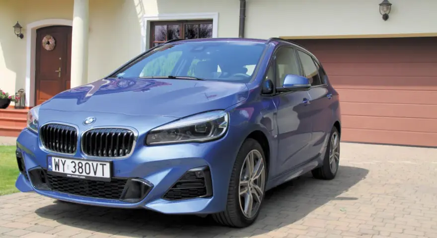 Niebieskie BMW 218i zaparkowane przed domem z brązową bramą garażową.