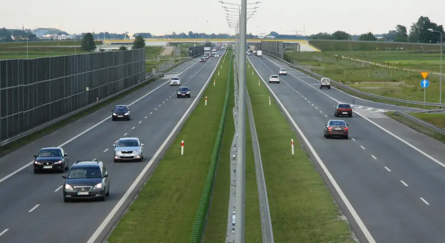 Na tej drodze szybkiego ruchu ruch jest umiarkowany. Widać kilka samochodów osobowych i jeden van. Po lewej stronie drogi znajduje się wysoki ekran akustyczny.