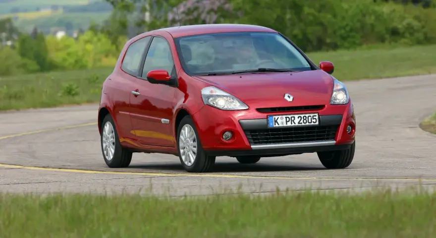 Czerwony Clio 3 na zakręcie drogi, otoczony zielenią.