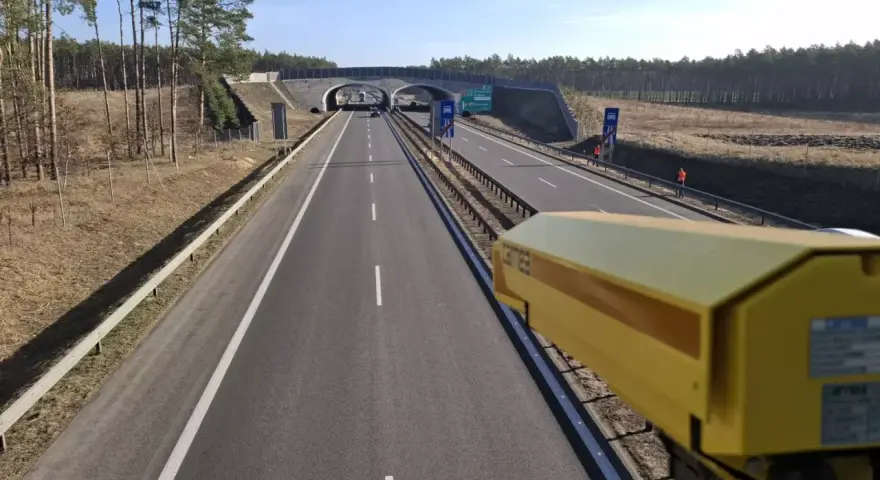 Żółta kamera monitoruje odcinek pomiarowy prędkości na S8. Droga prowadzi przez las, pod wiaduktem.