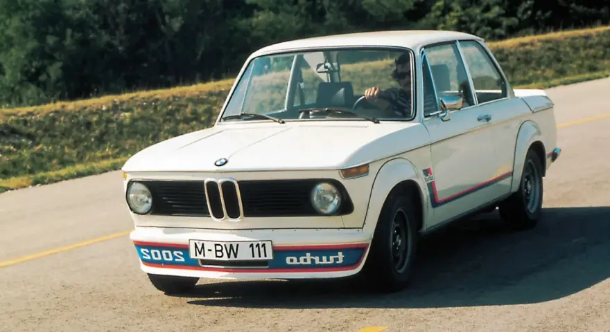 Białe BMW E20 z kolorowymi pasami i napisem "turbo" na błotniku, z kierowcą za kierownicą, jedzie po asfaltowej drodze.